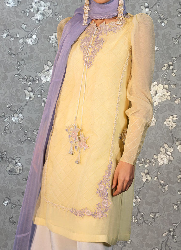 A person wearing a yellow dress with purple accents and a purple scarf, standing against a gray floral background.
