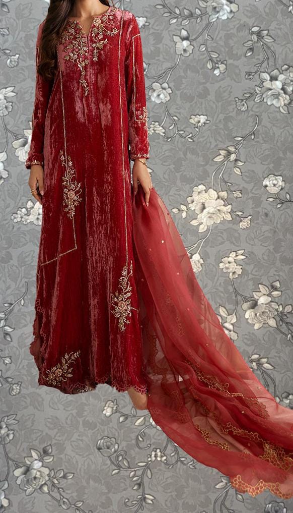 A woman wearing a long, red velvet dress with intricate embroidery stands against a gray floral background.