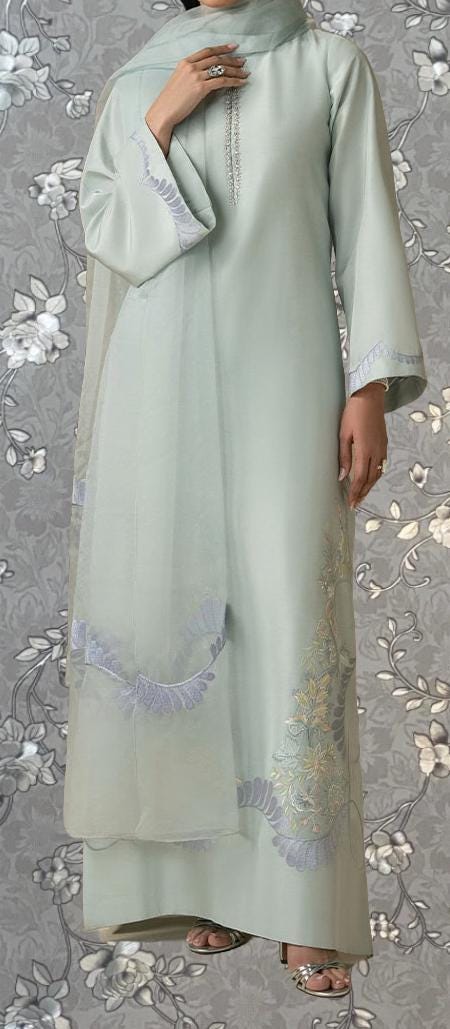 A woman wearing a light green dress with intricate embroidered designs on the sleeves and skirt, standing against a gray floral background.