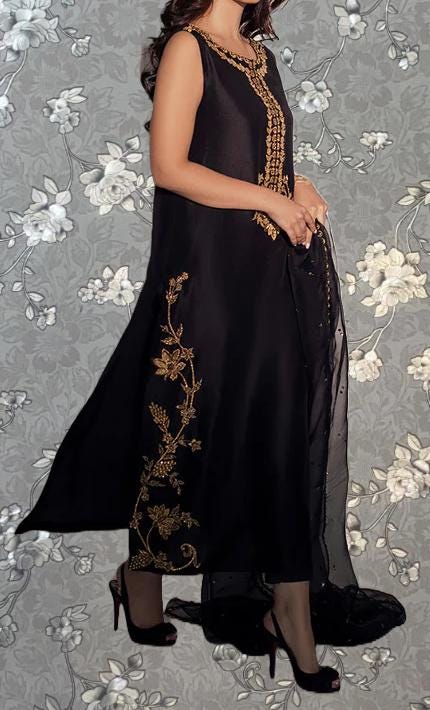 A woman wearing a black dress with gold embroidery stands against a gray floral background.