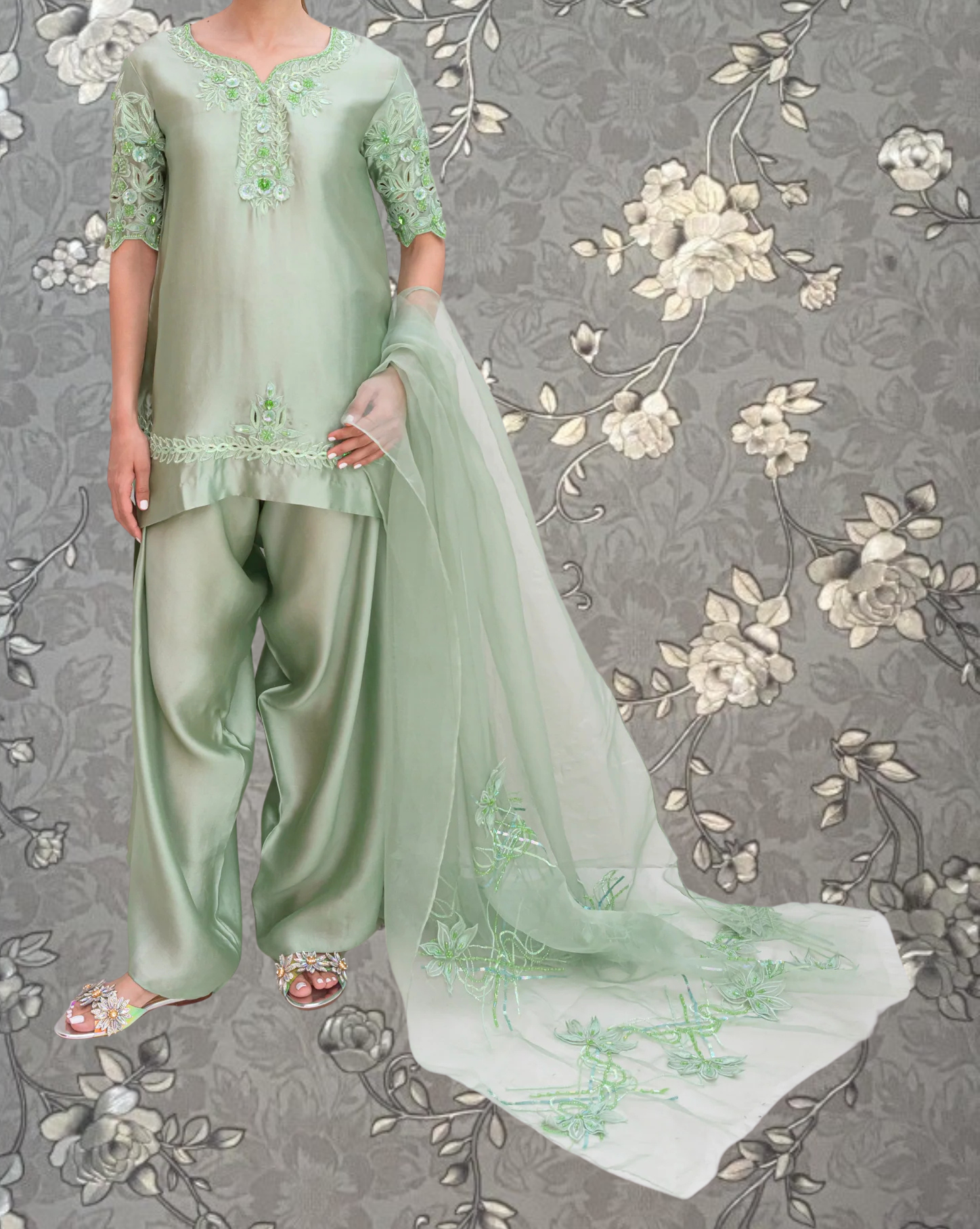 A person wearing a mint green dress with green floral embroidery stands against a gray floral background.
