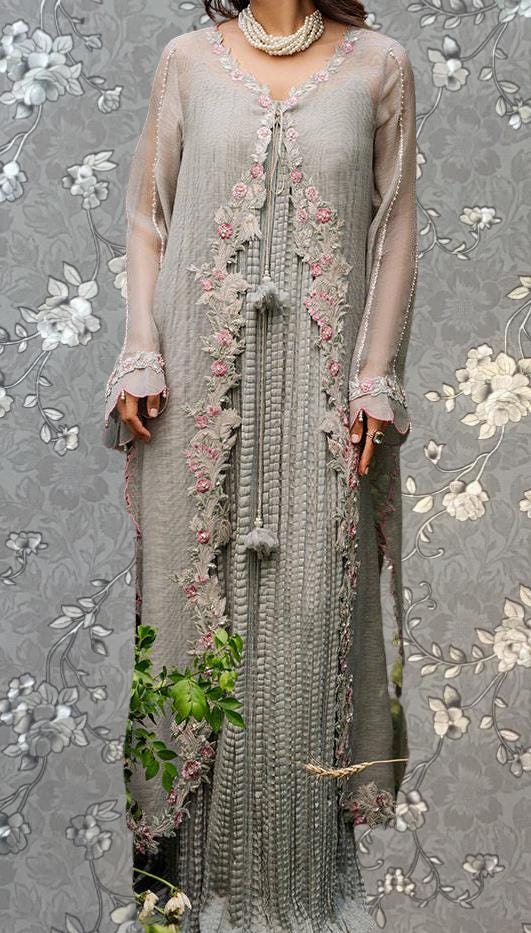 A woman wearing a long, gray dress with intricate floral embroidery and tassels, standing against a gray floral background.