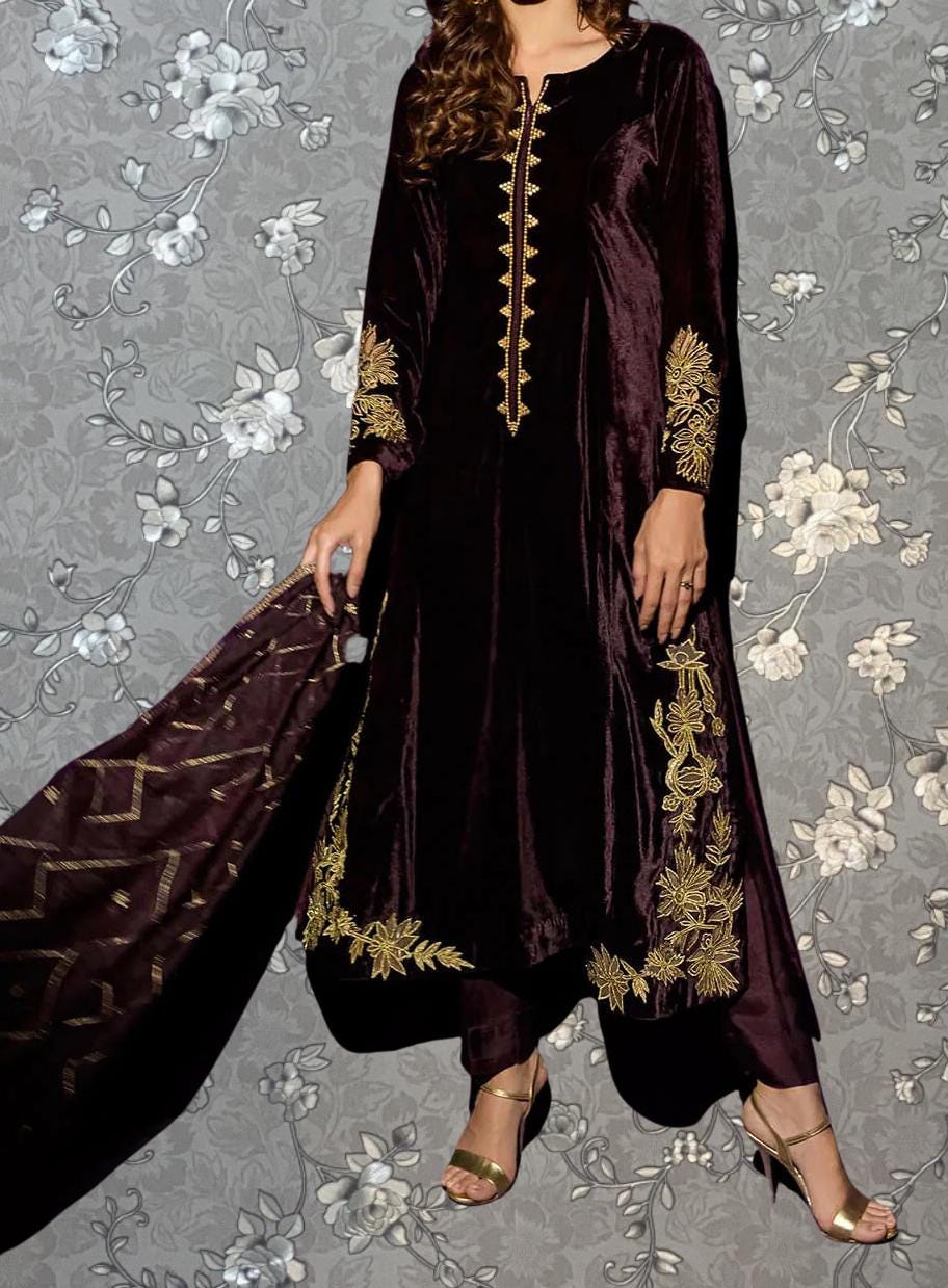 A woman wearing a long, burgundy velvet dress with gold embroidery stands against a gray floral background.