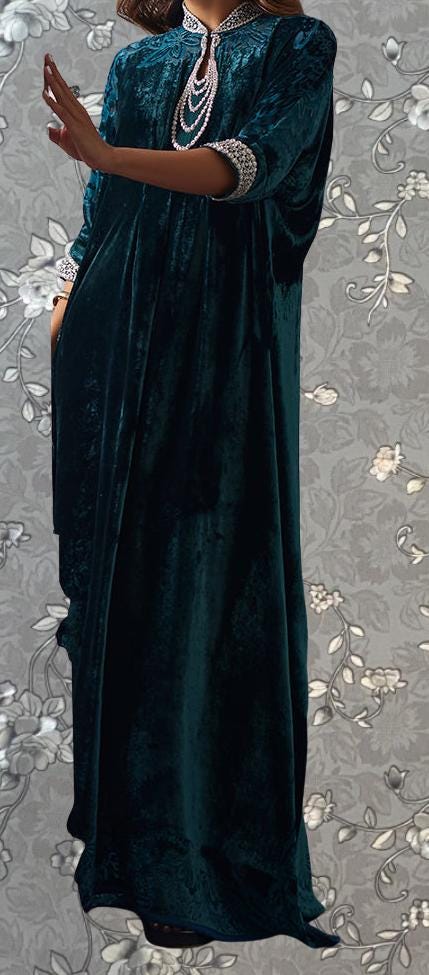 A woman wearing a long, dark green velvet dress with a high collar and sleeves, standing against a gray floral background.