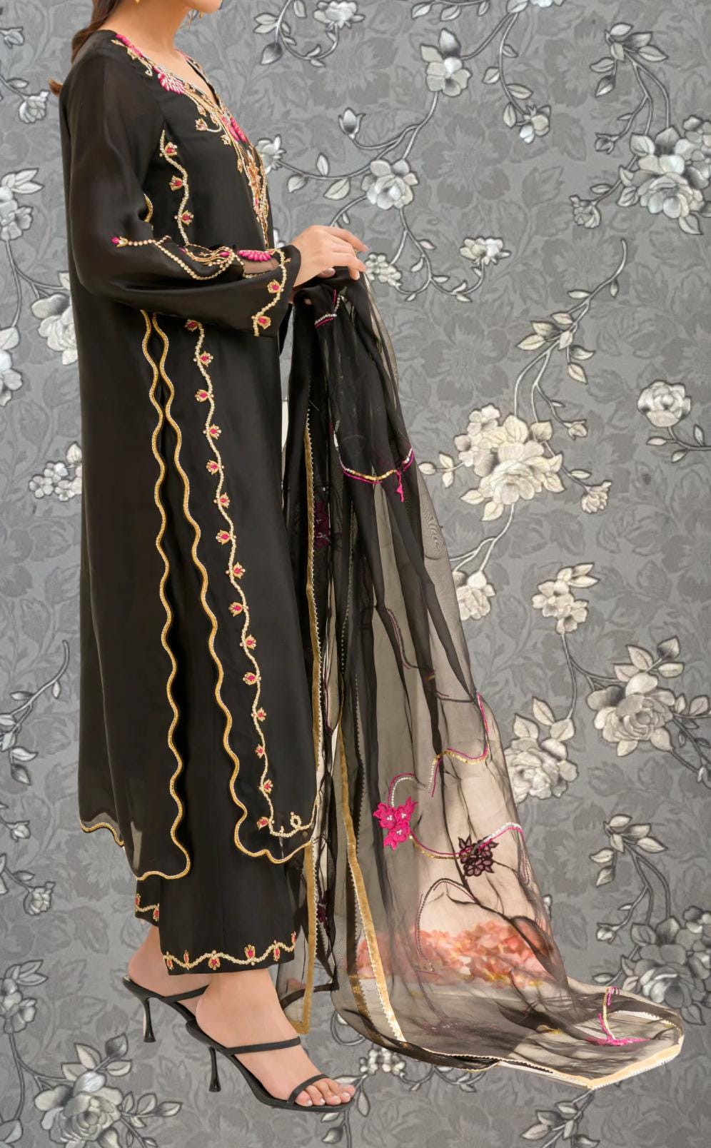 A woman wearing a long black dress with gold embroidery and a matching black shawl draped over her shoulders, standing against a gray floral background.