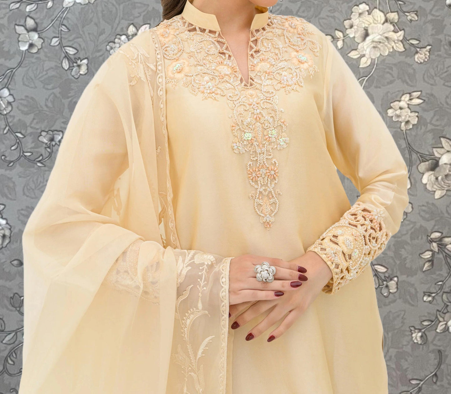A woman wearing a yellow dress with intricate embroidery and lace details, posing against a gray floral background.