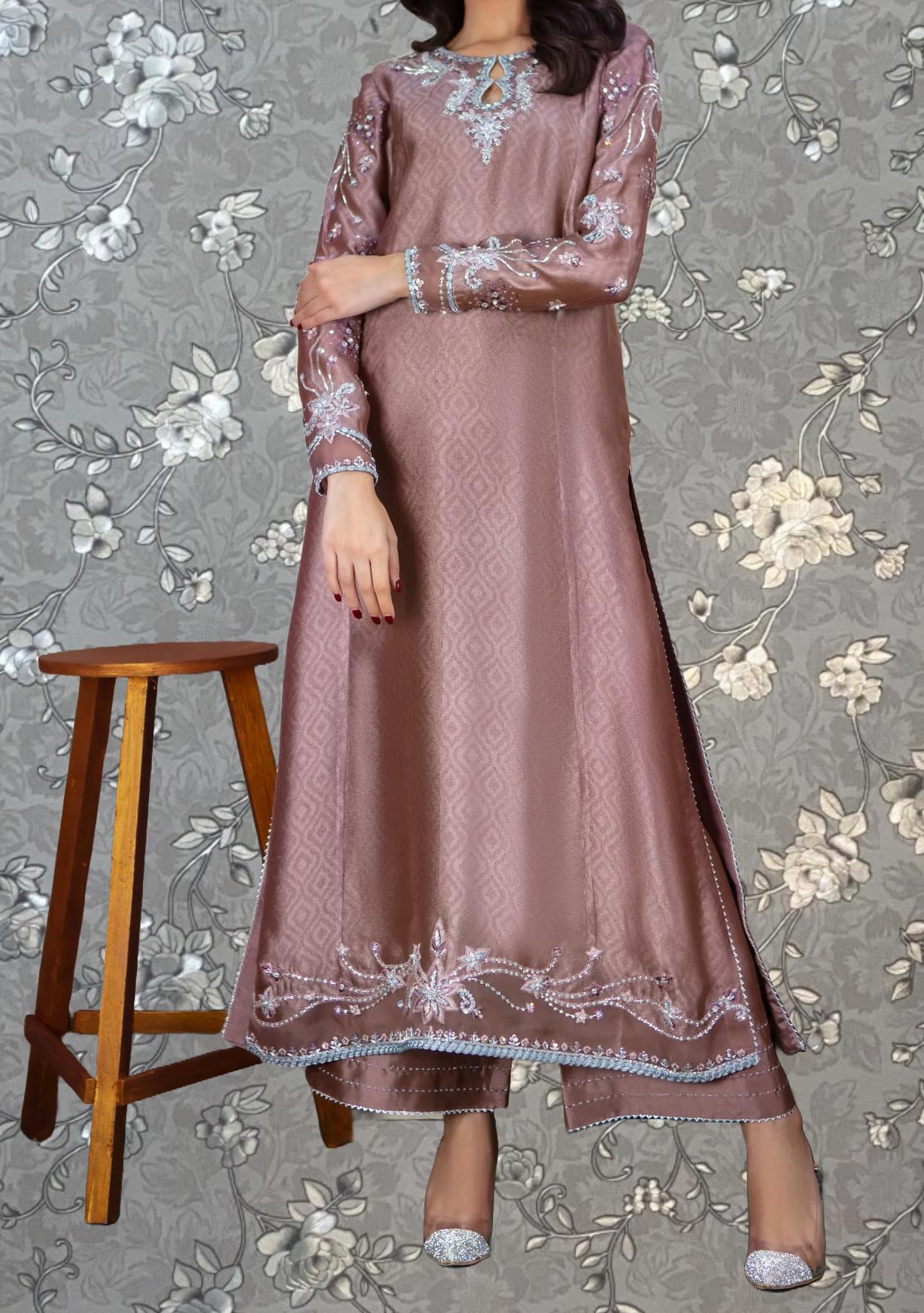 A woman wearing a long, pink dress with intricate embroidery stands in front of a floral patterned background.