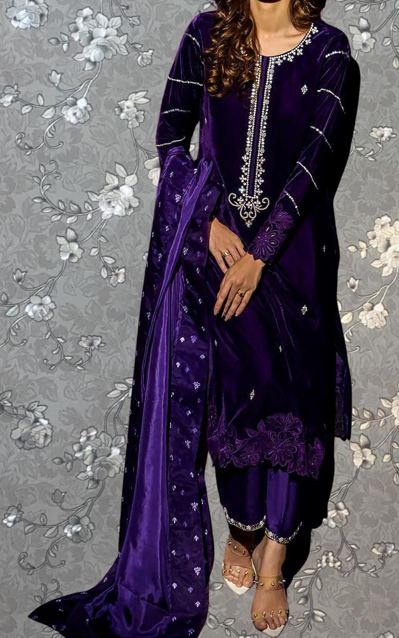 A woman wearing a purple velvet dress with intricate embroidery and a matching purple shawl stands against a gray floral background.
