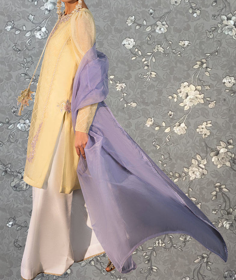 A woman wearing a yellow and purple dress stands against a gray floral background.