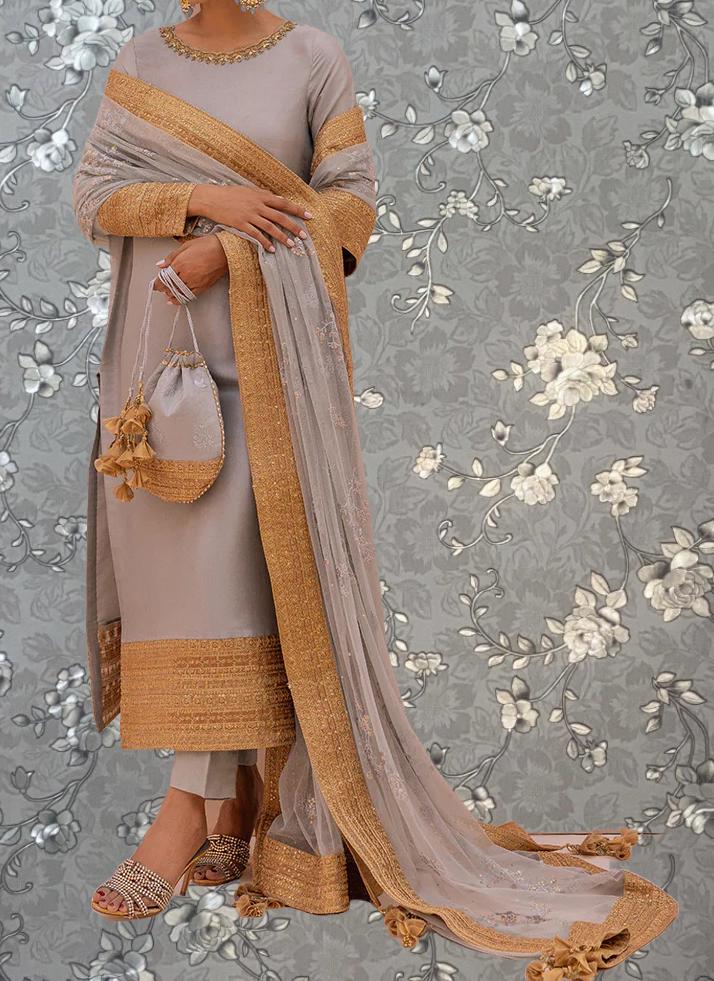 A woman wearing a gray and gold dress with a long, flowing skirt and holding a small purse stands against a gray floral background.