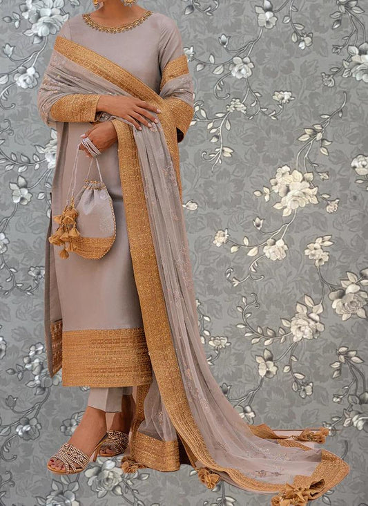 A woman wearing a gray and gold dress with a long, flowing skirt and holding a small purse stands against a gray floral background.