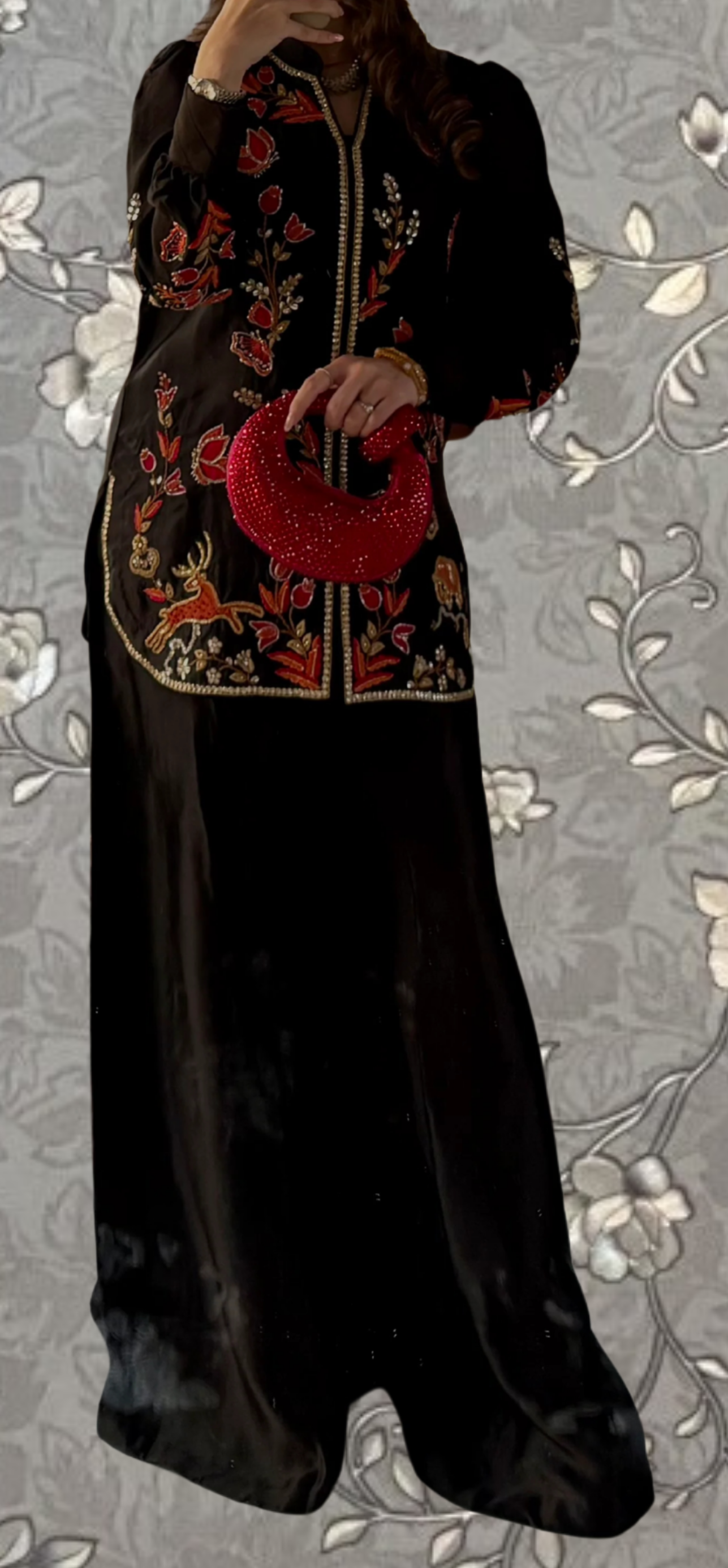 A person wearing a long black dress with red embroidered patterns and holding a red clutch purse.