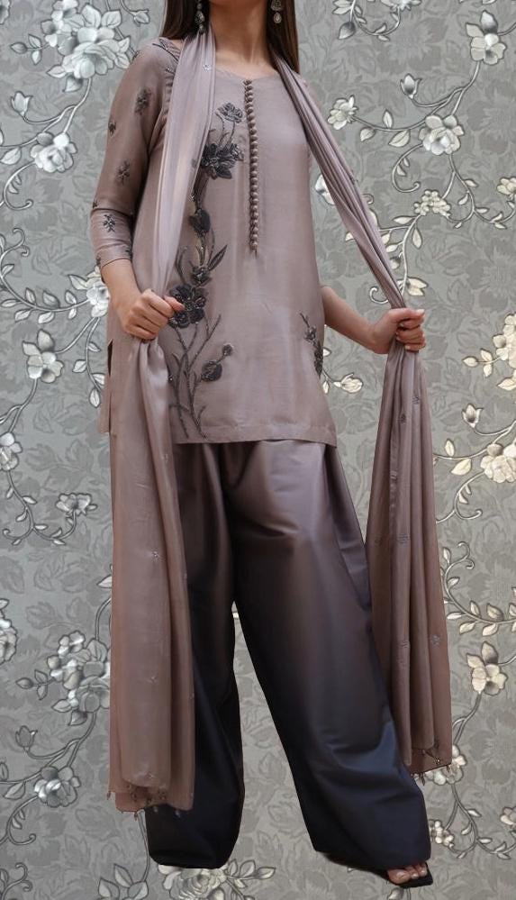A woman wearing a grey dress with floral embroidery and a matching grey shawl is standing against a gray floral background.