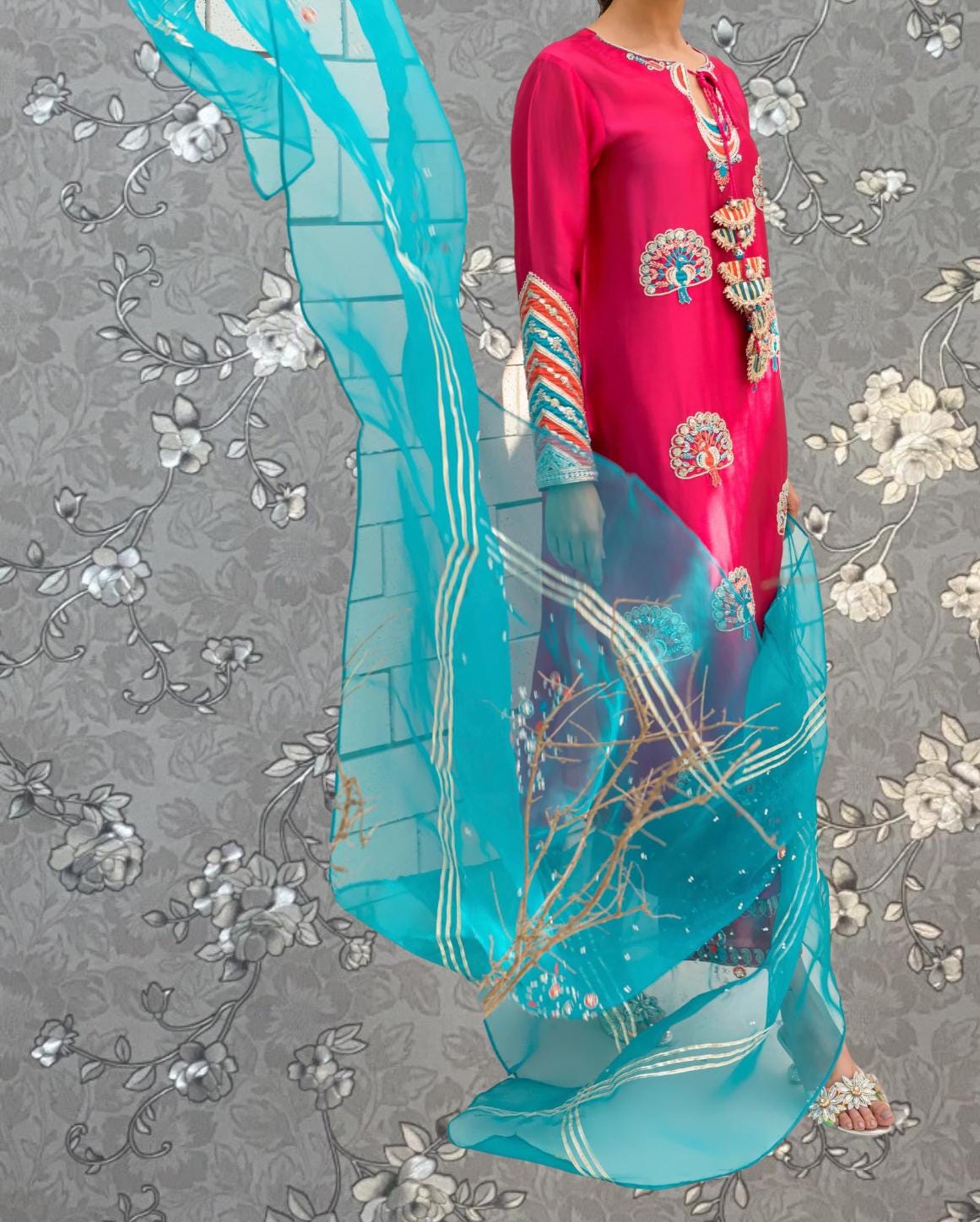 A person wearing a vibrant pink dress with intricate floral designs and a matching turquoise shawl stands against a gray floral background.