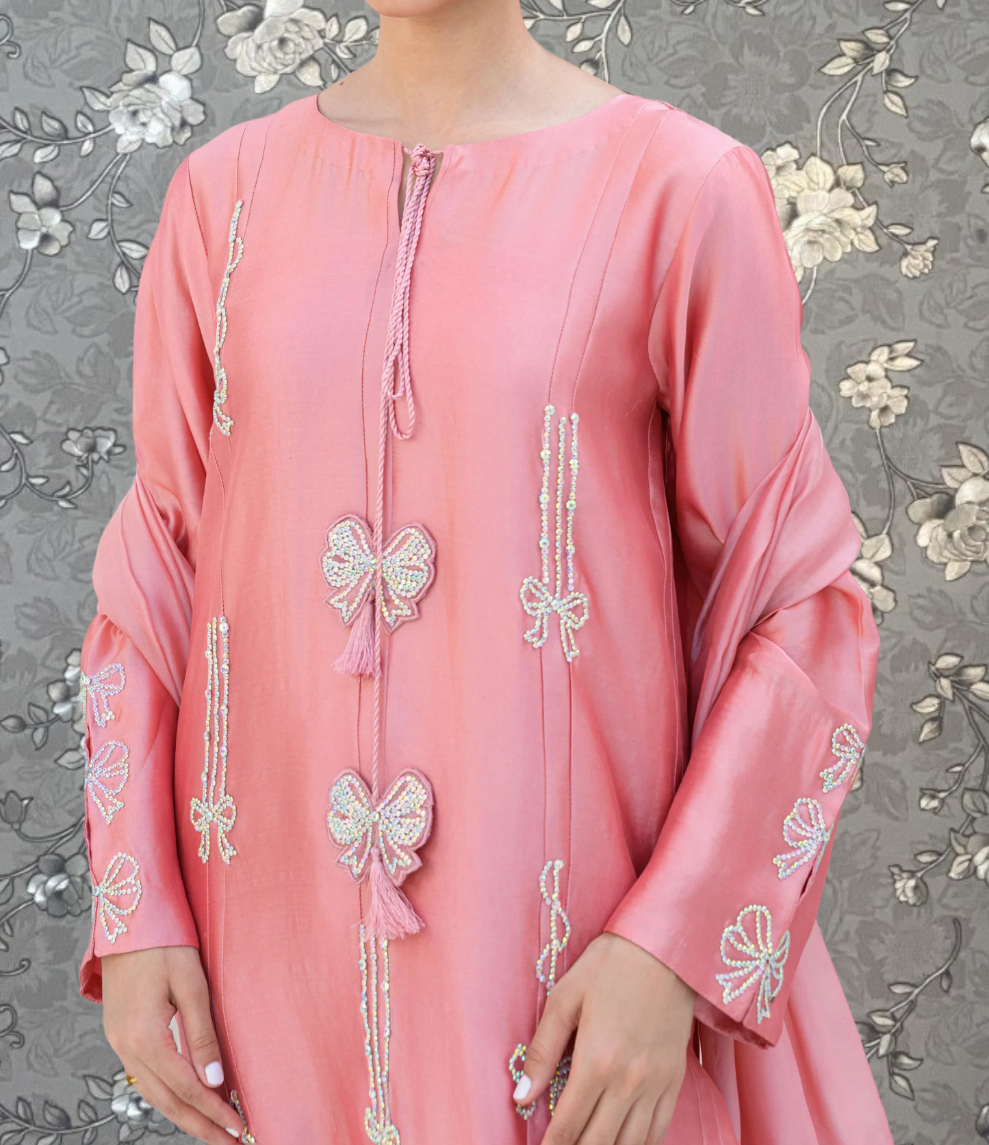 A woman wearing a pink dress with intricate embroidered designs, including bows and floral patterns, stands against a gray floral background.