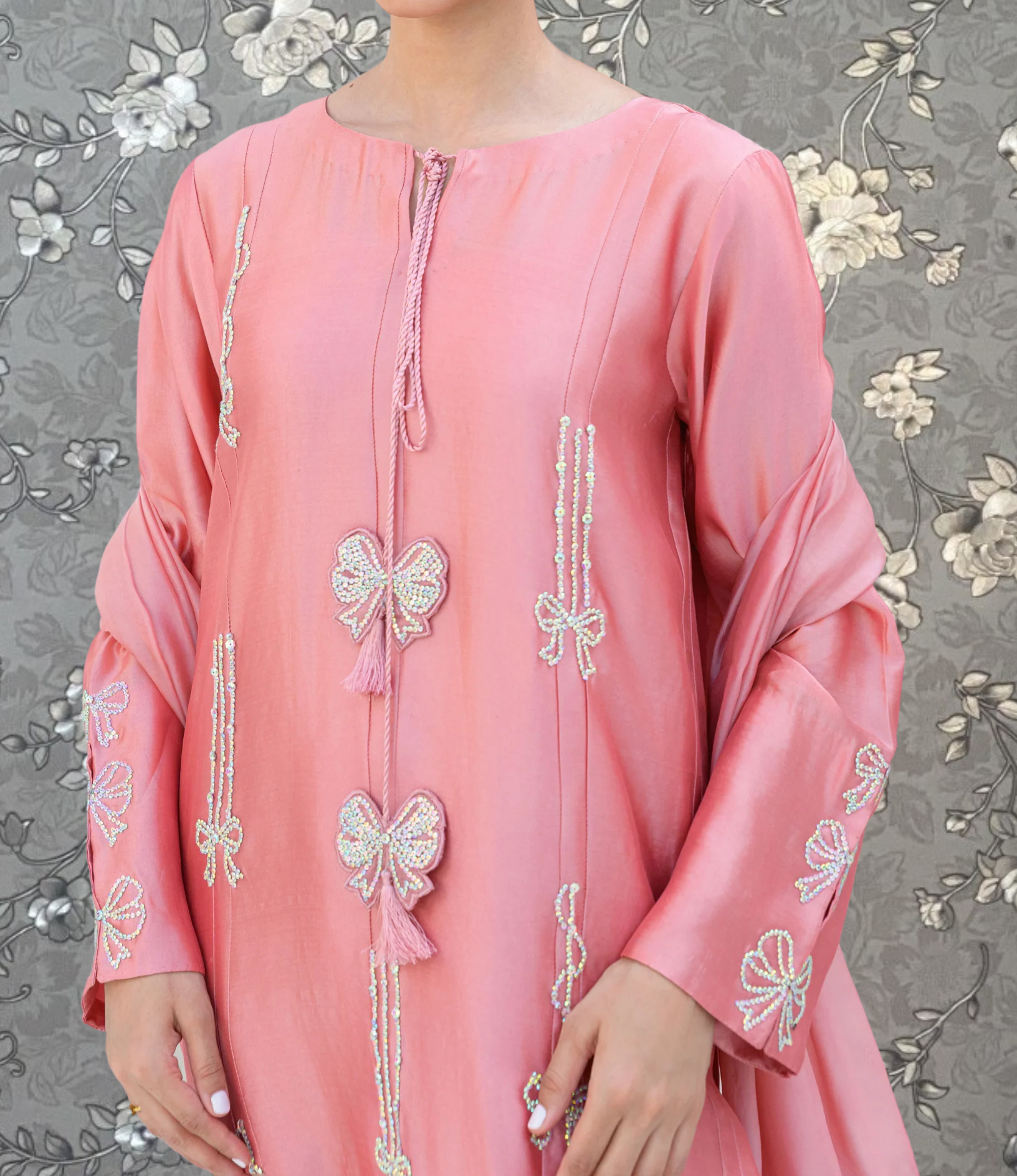 A woman wearing a pink dress with intricate embroidered designs, including bows and floral patterns, stands against a gray floral background.