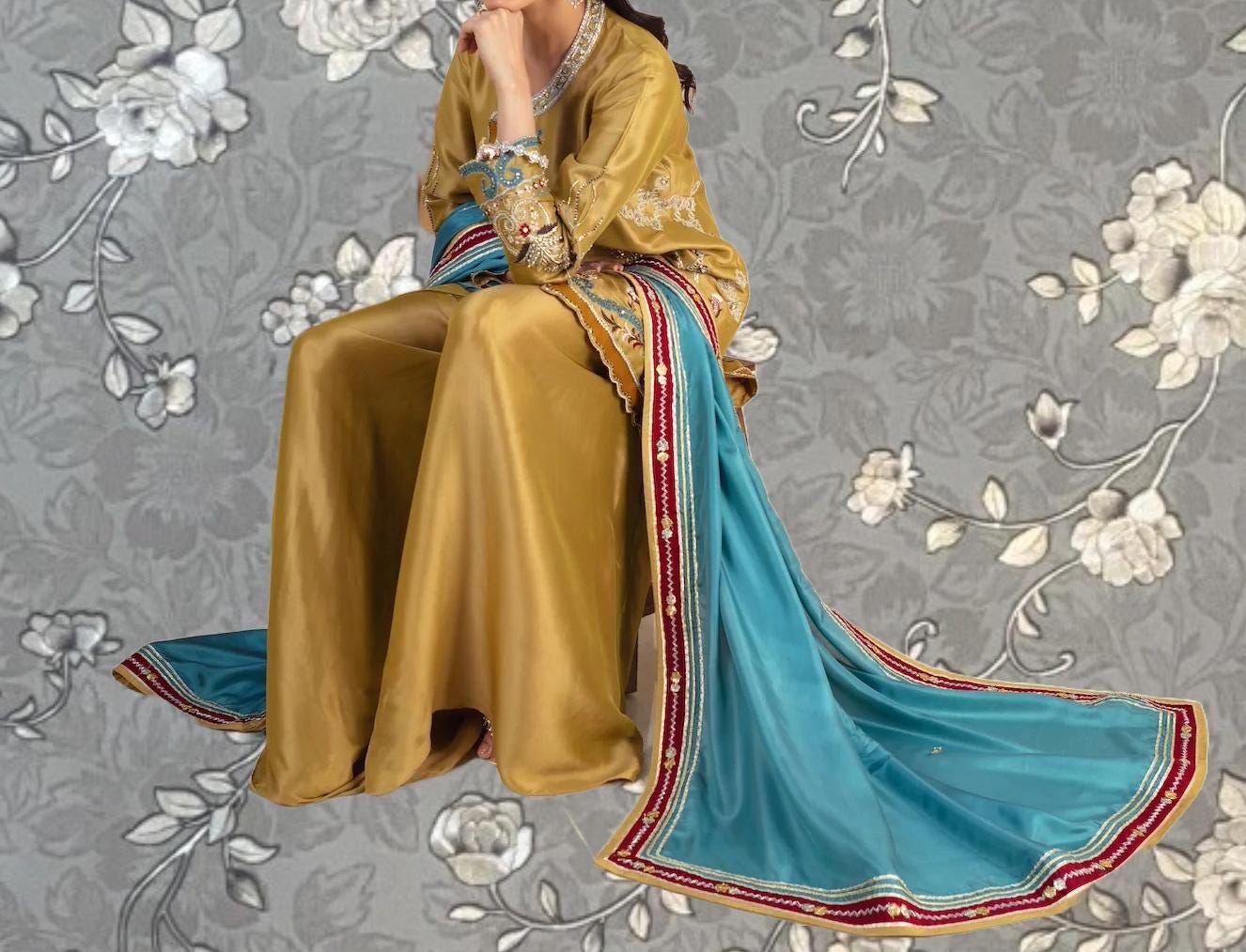 A woman wearing a gold and blue dress with a long train, sitting on a chair against a gray floral background.