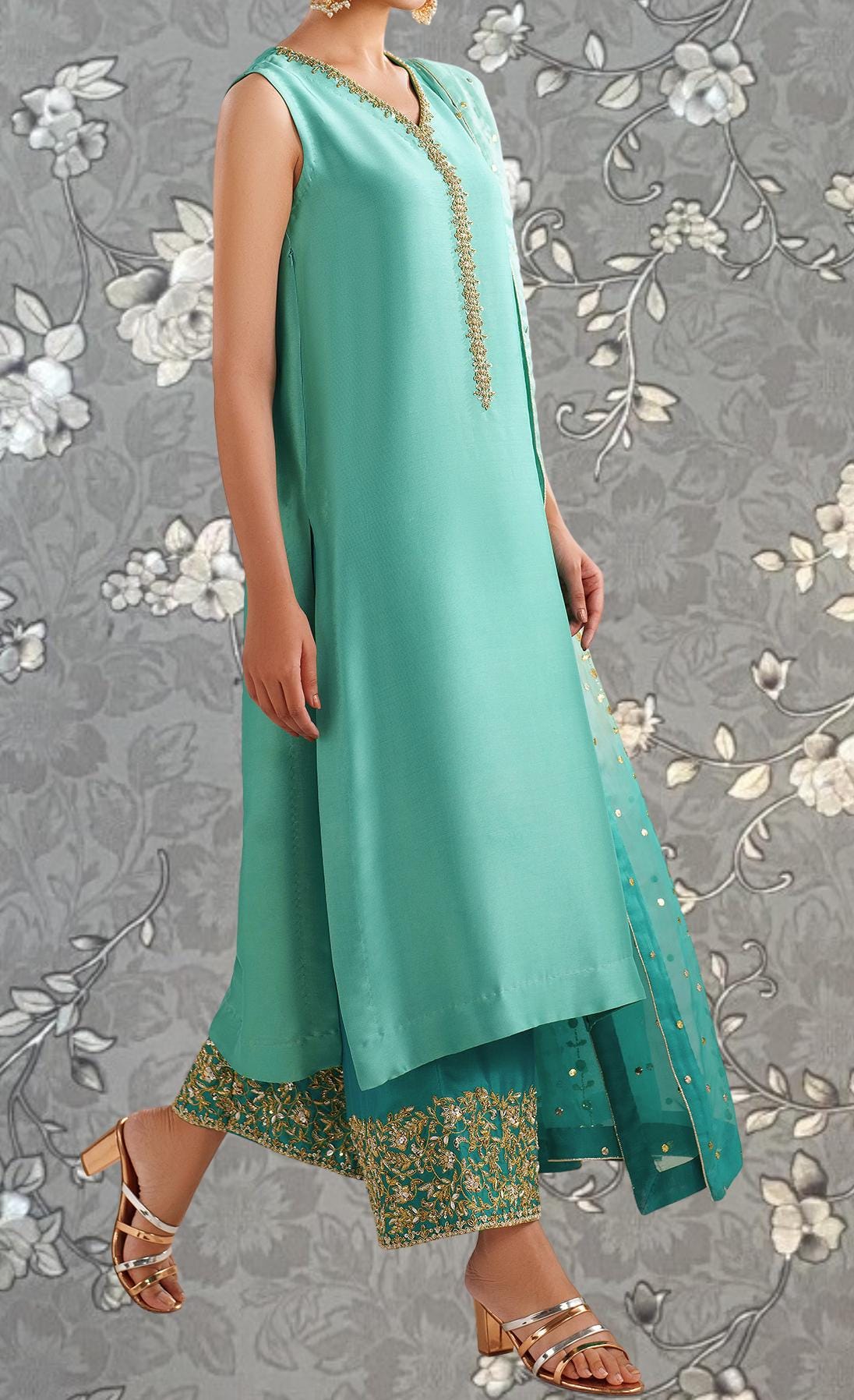 A woman wearing a turquoise dress with gold embroidery and gold heels is standing against a gray floral background.