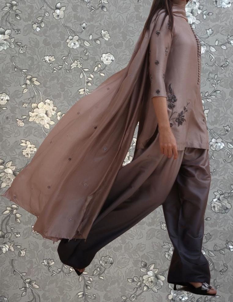 A woman wearing a long, flowing brown dress with floral embroidery is walking against a gray floral background.
