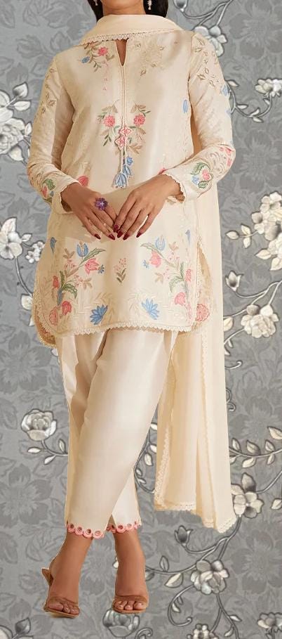 A woman wearing a cream-colored dress with floral embroidery stands against a gray floral background.