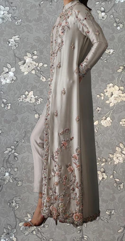 A woman wearing a long, gray dress with intricate floral embroidery stands against a gray floral background.