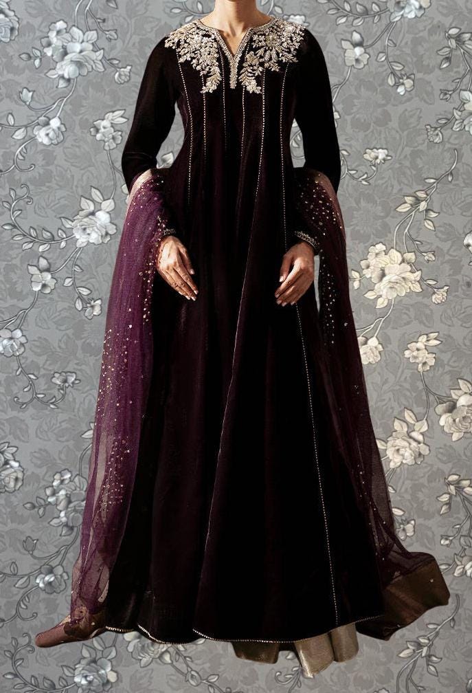 A woman wearing a long, dark purple dress with a high collar and long sleeves, standing against a gray floral background.