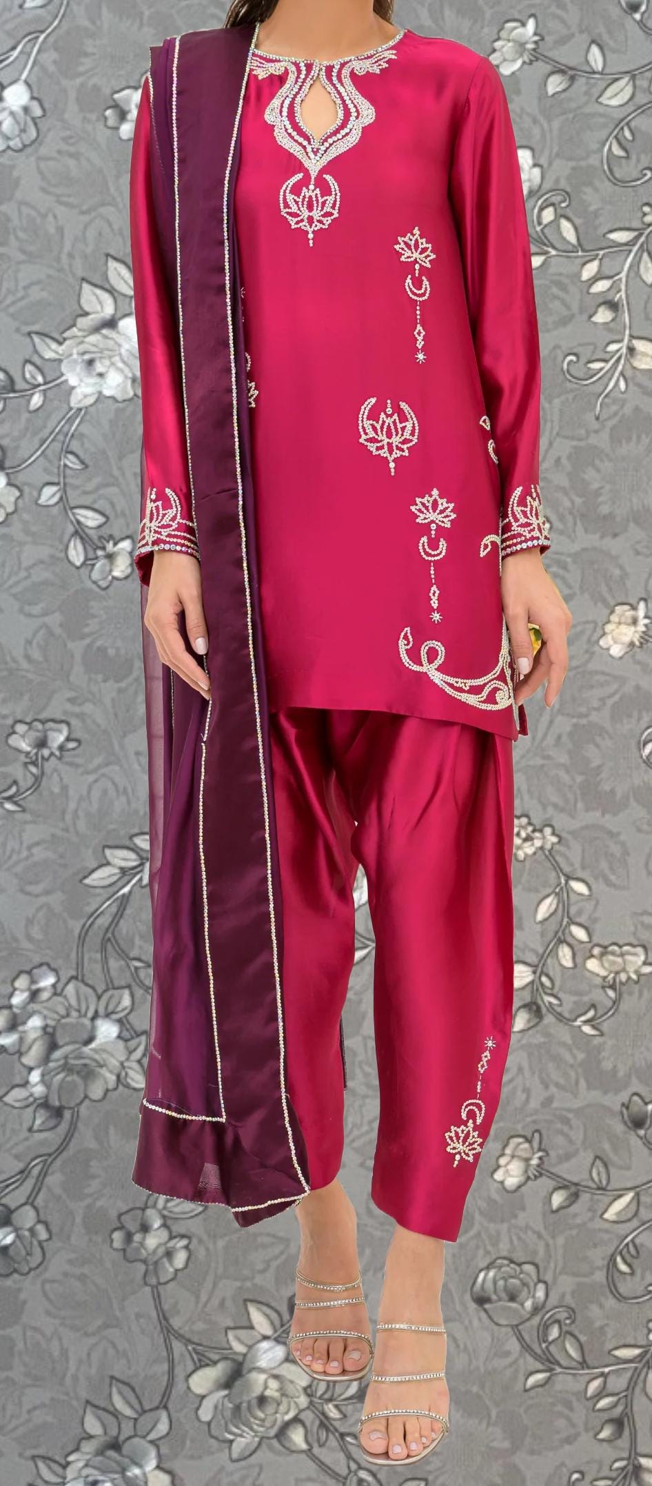 A woman wearing a red and purple outfit with intricate embroidery, standing against a gray floral background.