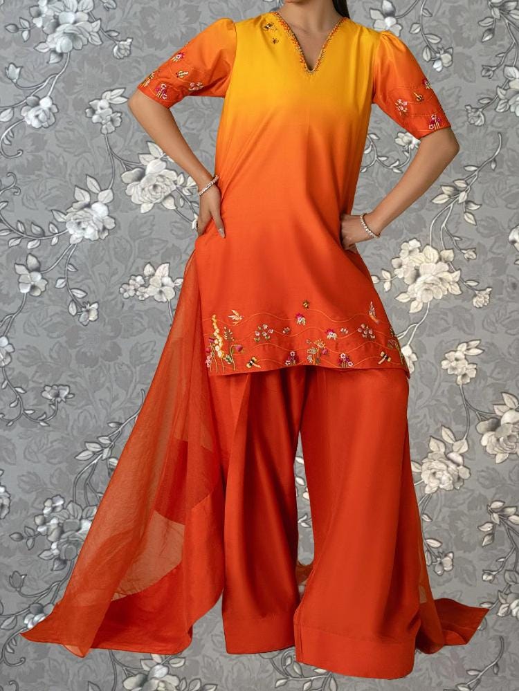 A woman wearing a vibrant orange and yellow dress with floral embroidery, standing against a gray floral background.