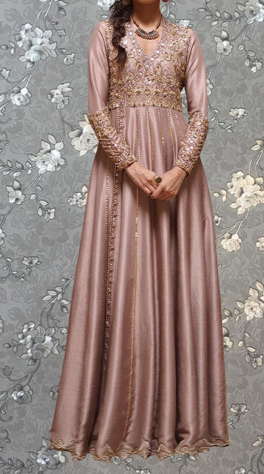 A woman wearing a long, pink dress with gold embroidery and a high neckline stands against a gray floral background.