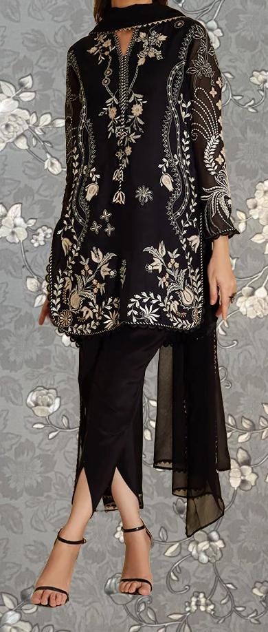 A woman wearing a black and white patterned dress with intricate embroidery, standing against a gray floral background.