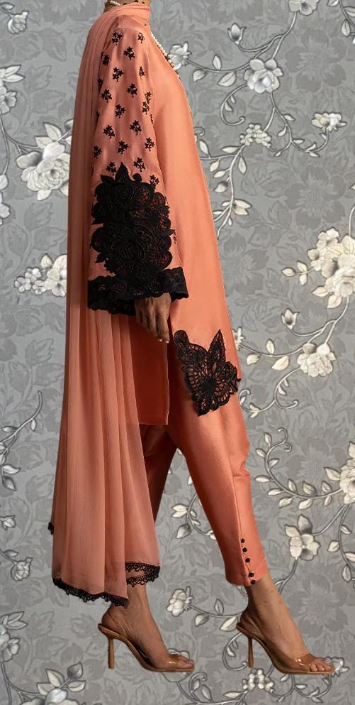 A person wearing a peach-colored dress with black floral embroidery and high heels, standing against a gray floral background.