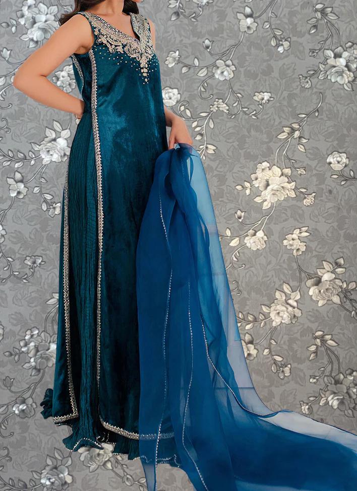 A woman wearing a long, blue dress with a matching blue shawl draped over her shoulders, standing against a gray floral background.