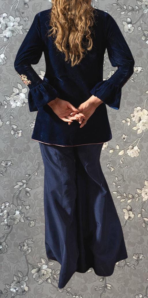 A woman with long, wavy hair wearing a navy blue velvet jacket and matching pants stands with her hands crossed in front of her.