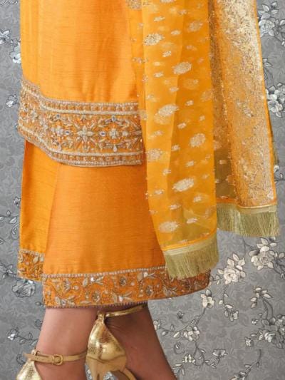a close-up view of a person wearing a vibrant yellow and orange outfit, including a skirt and a shawl. The outfit is adorned with intricate embroidery and embellishments, giving it a festive and elegant appearance. The person is also wearing gold high heels, which complement the overall color scheme of the outfit.