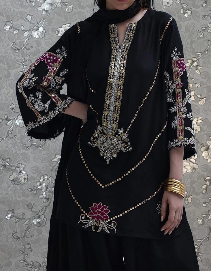 A woman wearing a black dress with intricate embroidery and gold accents, posing against a floral background.
