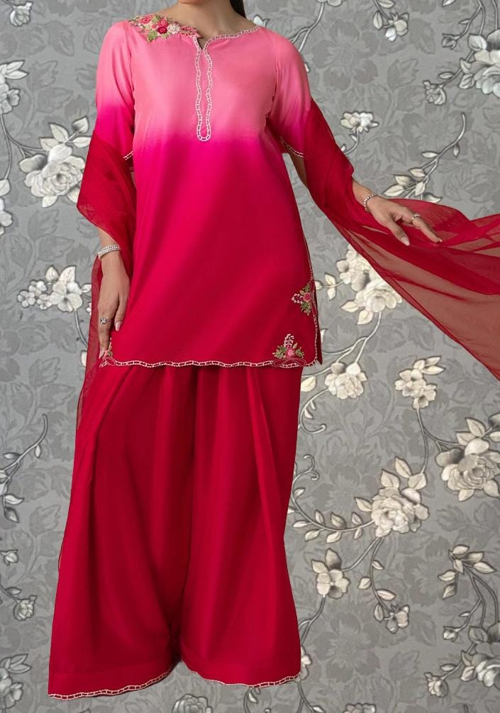 A woman wearing a pink and red dress with a floral pattern, standing against a gray floral background.