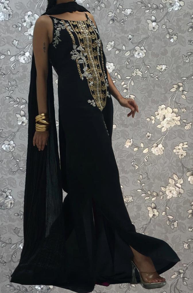 A woman wearing a long black dress with gold embroidery stands against a gray floral background.