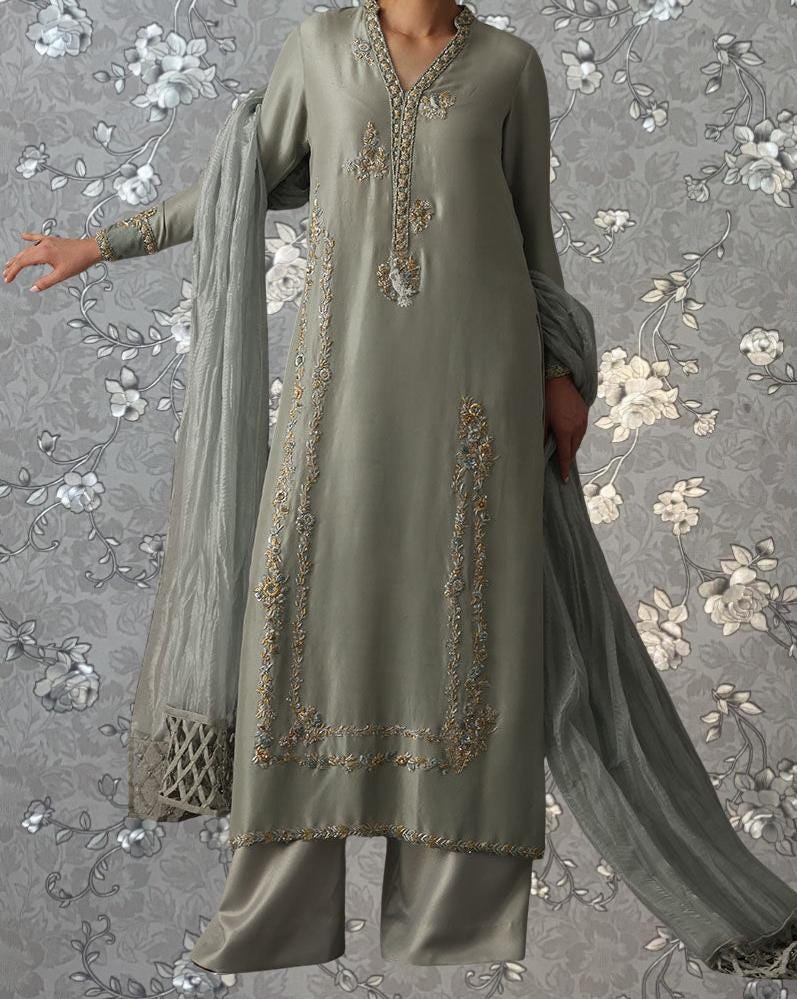 A person wearing a gray and gold embroidered dress with a matching gray shawl, standing against a gray floral background.