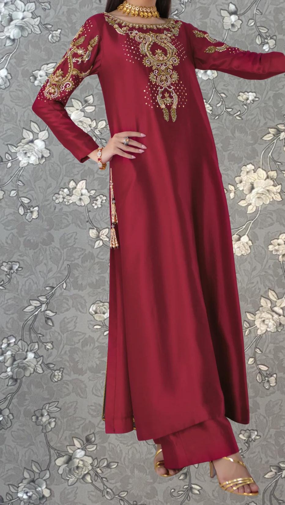 A woman wearing a long, burgundy dress with gold embroidery and tassels, posing against a gray floral background.