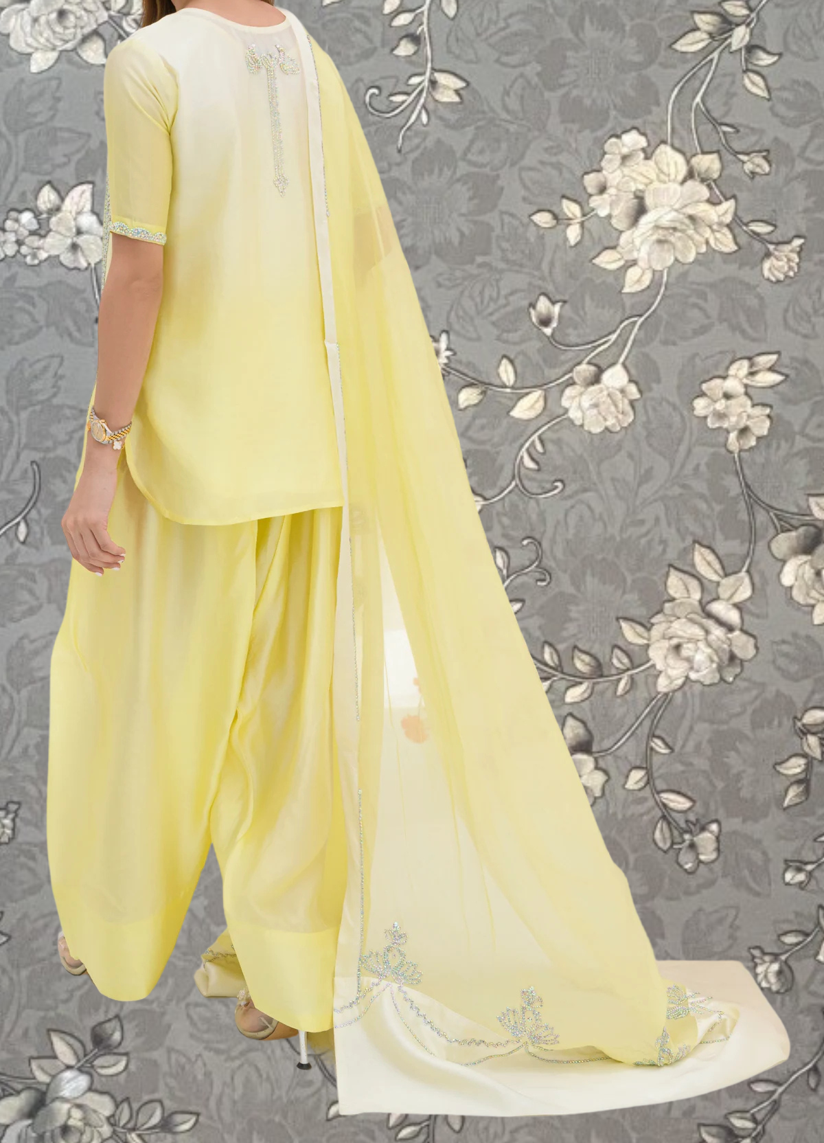 A person wearing a yellow dress with a long, flowing skirt and a matching yellow blouse stands against a gray floral background.