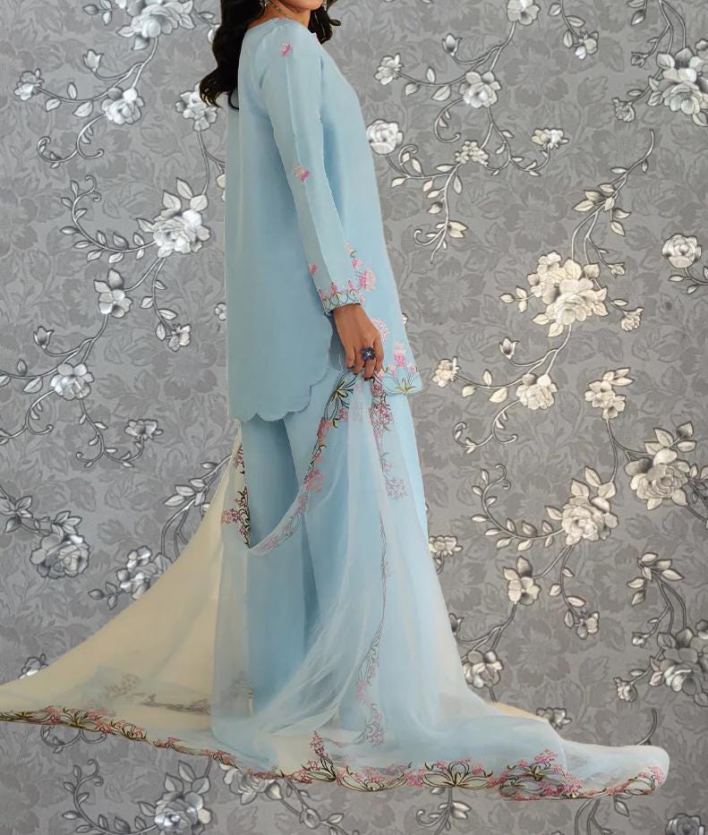 A woman wearing a light blue dress with floral embroidery stands against a gray floral background.