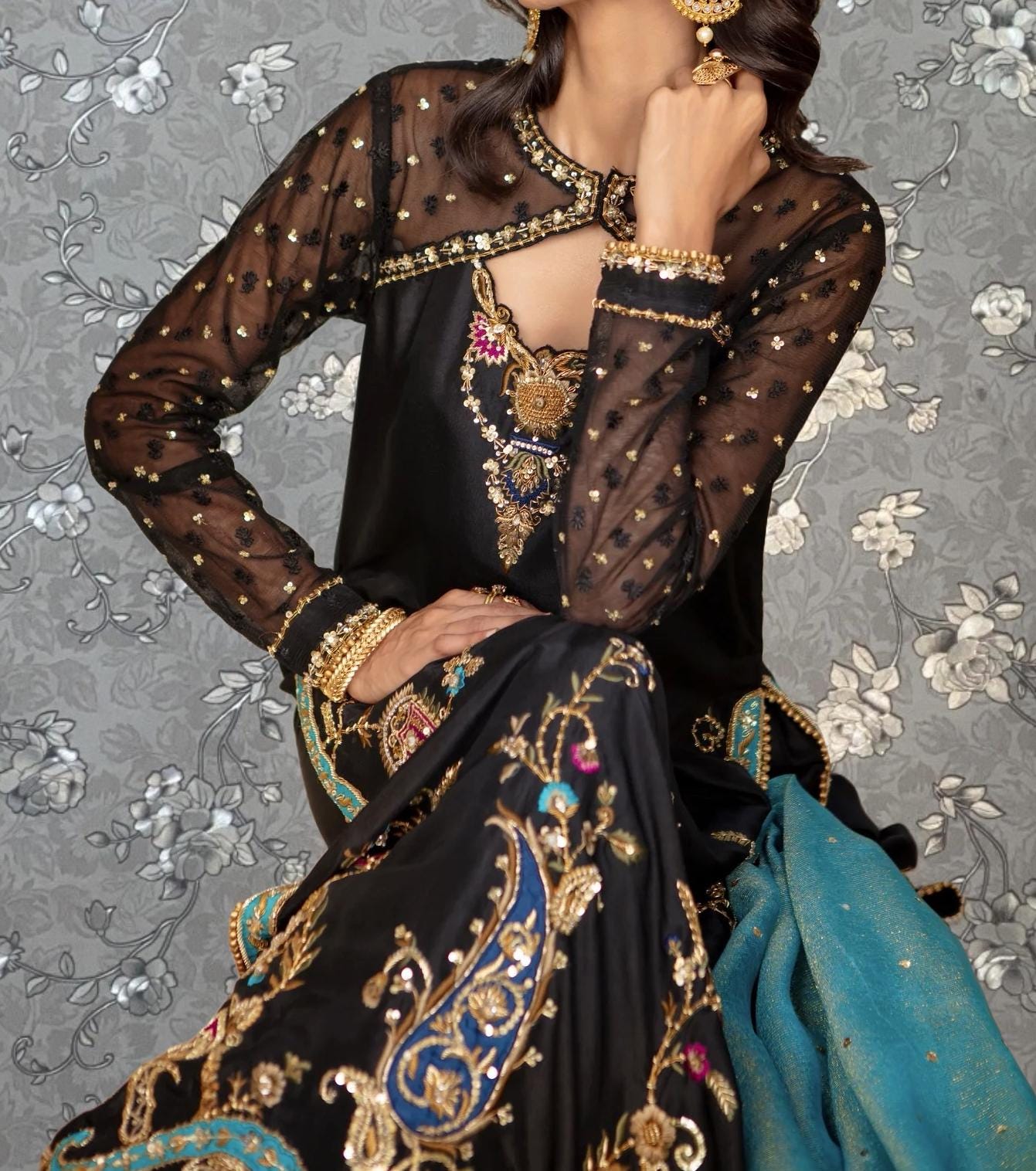A woman wearing an ornate, black and gold dress with intricate embroidery and jewelry poses against a gray floral background.