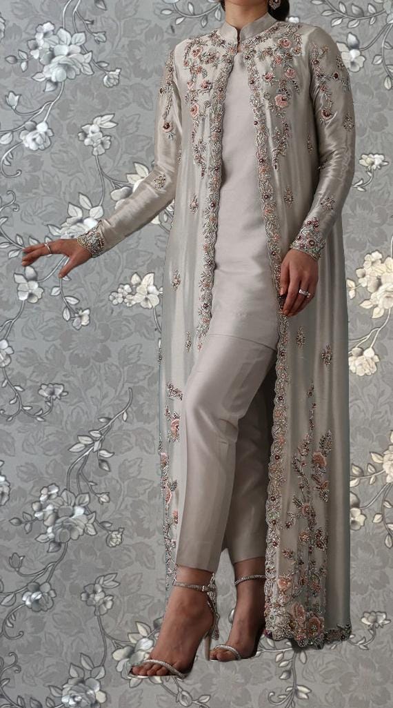 A woman wearing a long, beige dress with intricate floral embroidery stands against a gray floral background.