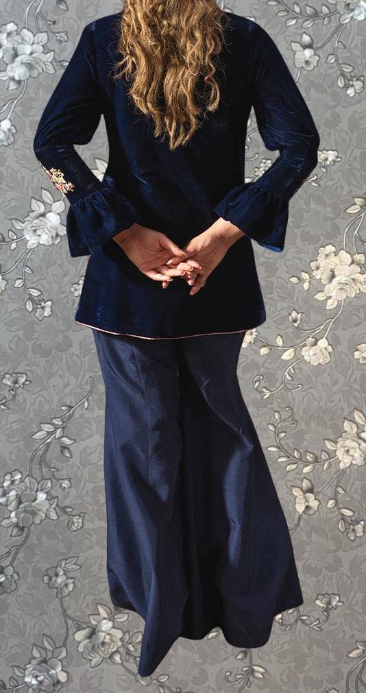 A woman wearing a navy blue velvet blazer and long navy blue pants stands with her hands clasped behind her back against a gray floral background.