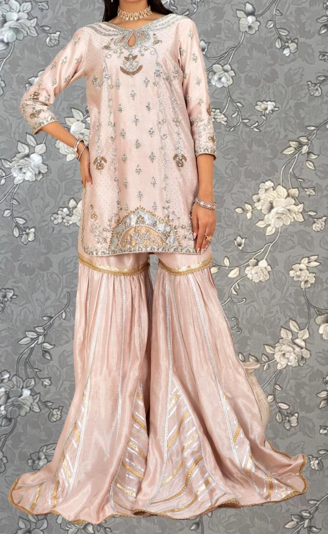 A woman wearing a pink and gold dress with intricate embroidery and a matching skirt, standing against a gray floral background.