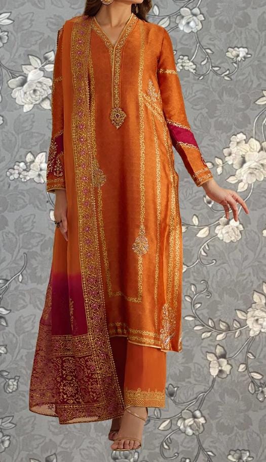 A woman wearing an orange and gold dress with intricate embroidery and patterns, standing against a gray floral background.