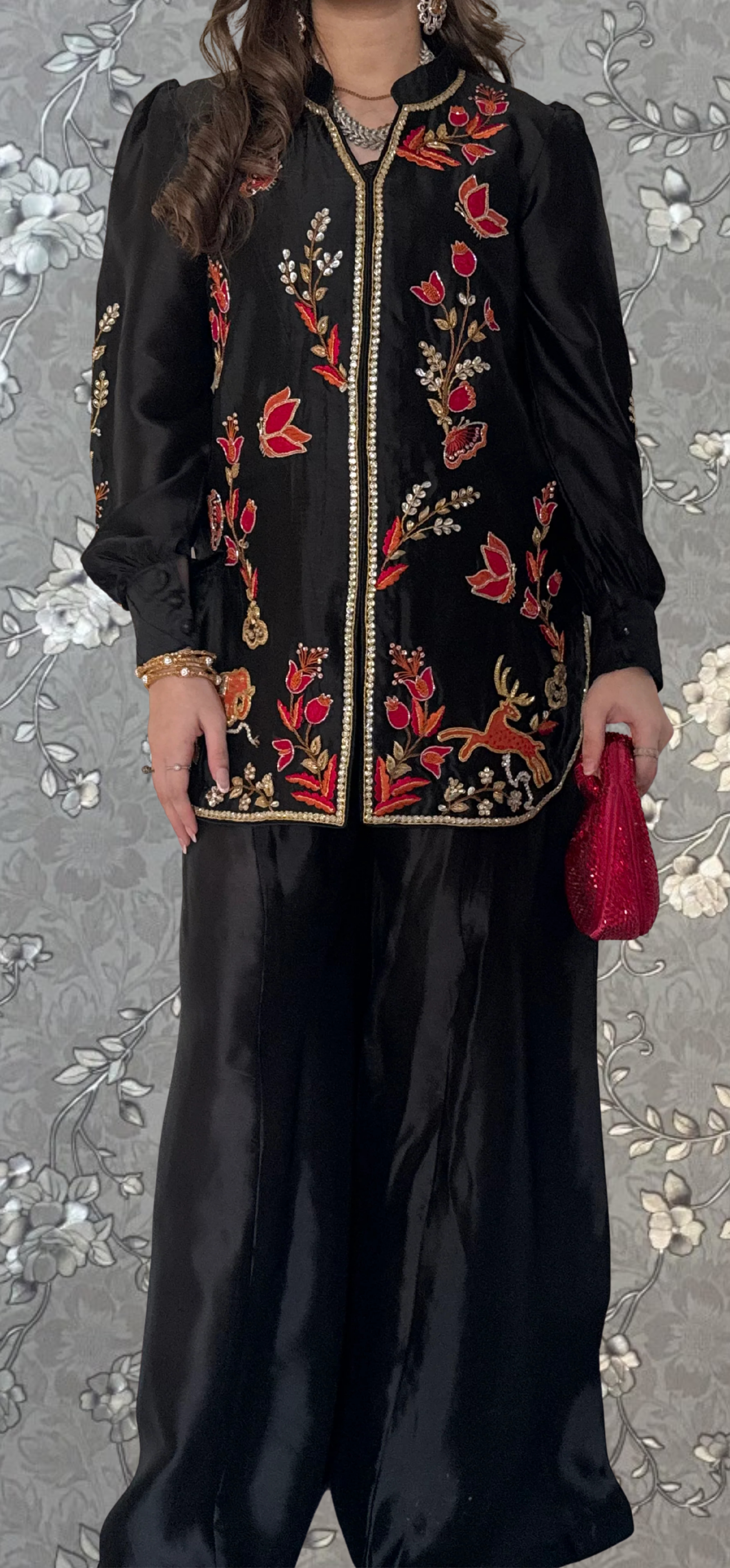 A woman wearing a black dress with red floral embroidery and holding a red clutch purse stands against a gray floral background.