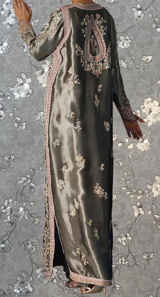a person wearing a long, gray dress with intricate embroidery and floral patterns. The dress has a high neckline and long sleeves, giving it a formal and elegant appearance. The person is standing against a gray floral background, which complements the overall design of the dress.