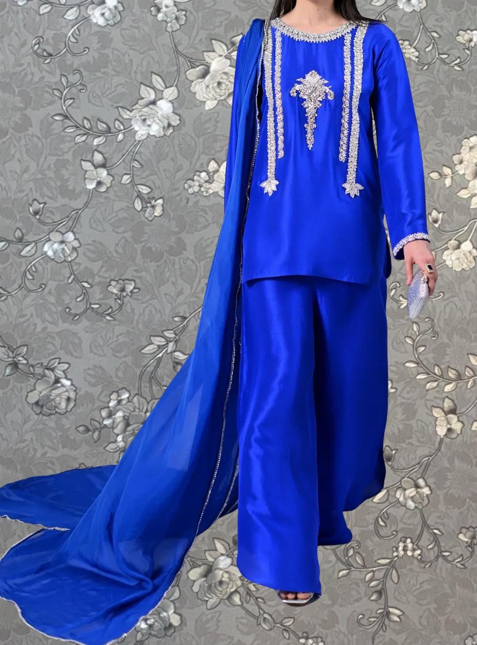 A woman wearing a blue dress with white embroidery and a long blue cape, standing against a gray floral background.