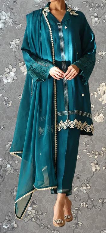 A person wearing a teal-colored dress with intricate embroidery and a matching shawl stands against a gray floral background.